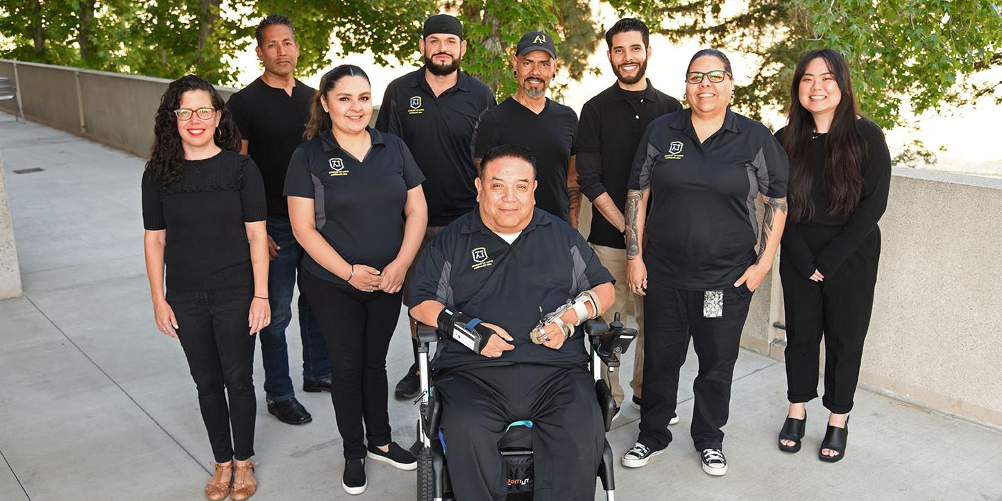 Nine people wearing matching shirts standing in a shady areas outdoors, smiling.