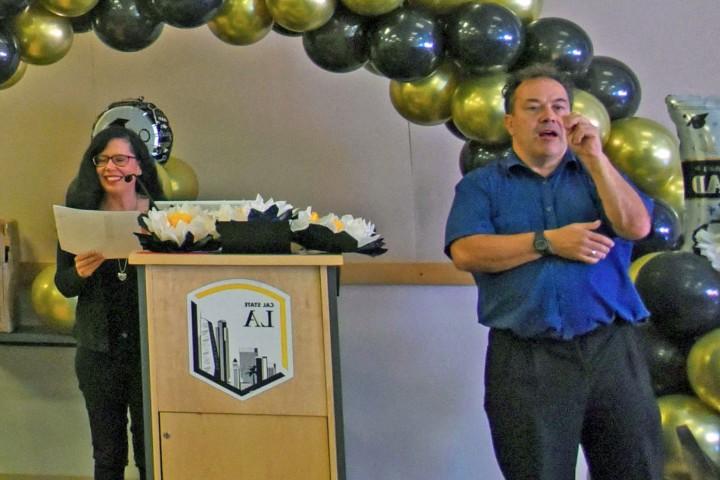 Sign language interpreter stands and signs in front of presenter at a podium.